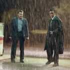 Zwei nasse Männer in Anzügen stehen im Regen vor einer Tankstelle. Einer hält eine Waffe. Im Hintergrund steht „famous“.
