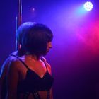 Eine Frau und ein Mann stehen Rücken an Rücken vor einer Stange. Die Bühne ist in blauem und rotem Licht beleuchtet.