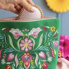 Ein grünes Täschchen mit bunten Blumenstickereien. Eine Hand hält eine kleine Bürste über dem offenen Reißverschluss.