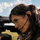 Eine Frau mit Headset zielt konzentriert durch ein Gewehrvisier. Hintergrund: blauer Himmel mit Wolken.