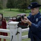 Ein Mann in Polizeiuniform zielführend mit einer Pistole. Eine Frau im roten Mantel steht daneben. Im Hintergrund Schafweide.