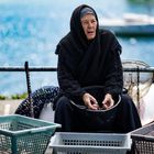 Eine ältere Frau in schwarzer Kleidung sitzt am Wasser, hält Muscheln in den Händen und umgeben von einigen Körben.
