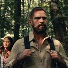 Ein Mann mit Bart und Rucksack geht durch einen Wald, gefolgt von einer Frau. Sie trägt ein kariertes Hemd.