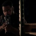 Eine Person zielt mit einem Gewehr, teilweise verdeckt durch ein Regal, in einem dunklen Raum.