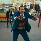 Ein Mann im blauen Anzug mit Timer "09:59:10" auf der Weste. Menschenmenge im Hintergrund, er sieht besorgt aus.