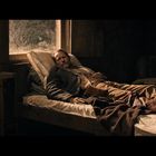 Ein Mann liegt in einem dunklen Raum auf einem Bett und blickt nachdenklich, Licht fällt durch ein Fenster.