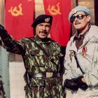 Zwei Männer in Militäruniformen, einer zeigt nach vorne. Rote Fahnen mit Hammer-und-Sichel-Symbol im Hintergrund.