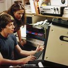 Zwei Jugendliche schauen auf einen alten Computerbildschirm. Ein Junge tippt auf der Tastatur, ein Mädchen lehnt sich über ihn.