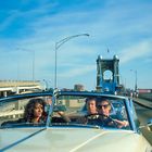 Drei Personen in einem offenen Auto; im Hintergrund eine Brücke und ein klarer Himmel.