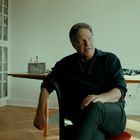 Ein Mann sitzt in einem Raum mit Bücherregalen und einem Tisch. Fenster und Heizung im Hintergrund.