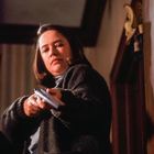 Eine Frau in dunkler Kleidung hält ein Gewehr. Sie blickt ernst und konzentriert in den Raum.