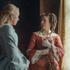 Zwei Frauen in viktorianischer Kleidung, unterhalten sich in einem elegant eingerichteten Raum neben einem Fenster.