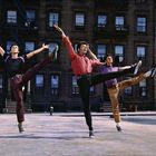 Drei Männer tanzen synchron vor einem Backstein-Gebäude. Ihre Arme und Beine sind in ausdrucksvollen Posen gestreckt.