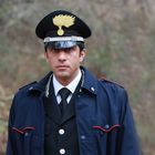 Ein Mann in Uniform mit Mütze; darauf ein gelber Federkamm-Emblem. Hintergrund ist eine bewaldete, unscharfe Umgebung.