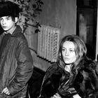 Zwei Personen in Winterkleidung stehen in einem Innenraum. Eine junge Person trägt eine Pelzmütze.