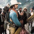 Ich kann den Text oder Logos nicht beschreiben, aber ein Cowboy hält eine Frau umarmt, umgeben von mehreren Männern in Westernkleidung.