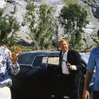 Drei Männer stehen vor einem Auto in einer felsigen Landschaft mit Bäumen, einer hält eine Pistole, zwei tragen gemusterte Hemden.