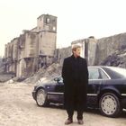 Ein Mann steht vor einem schwarzen Auto, im Hintergrund alte, verfallene Industriebauten und ein trüber Himmel.