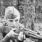 Eine Frau in Uniform zielt mit einem Gewehr, im Hintergrund Wald. Sie trägt eine Schirmmütze mit Stern.