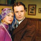 Ein Paar in eleganter Kleidung: Sie trägt einen lila Kopfschmuck mit Federn, er einen braunen Anzug in einem Raum mit Holzverkleidung.