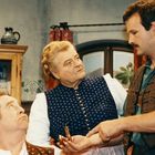 "Innst es dein Mütterlein" steht auf einer Gardine. Drei Menschen in traditioneller Kleidung, Frau hält Handgerät.