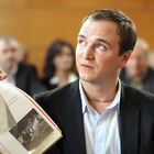 Eine Person im Anzug sitzt, schaut interessiert auf ein aufgeschlagenes Dokument mit zwei Fotos. Im Hintergrund sind unscharfe Zuschauer.