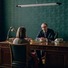 Ein Mann im Anzug sitzt an einem Schreibtisch und spricht mit einer Frau. Der Raum hat grüne Wände.