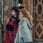 Zwei Frauen in historischen Kostümen, eine in Rot, die andere in Blau, in einem opulenten Raum mit Marmorelementen.