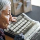 Eine Person mit grauen Haaren betrachtet ein Album mit Fotos oder Karten. Im Hintergrund unscharfe Kisten.