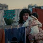 Eine Frau hält ein Baby im Arm. Im Hintergrund hängt bunte Wäsche an einer Leine, und Gebäude sind sichtbar.