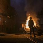 Eine Person steht vor einem brennenden Auto, das Feuer lodert aus der Motorhaube in einer dunklen, industriellen Umgebung.