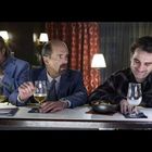 Drei Männer sitzen an einer Bar. Der mittlere Mann blickt ernst, während die anderen trinken. Im Hintergrund stehen Lampen.