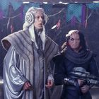 Zwei Fantasiefiguren in aufwendigen Kostümen; einer trägt eine Waffe. Bunte Dekorationen im Hintergrund.