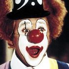 Ein Clown mit roter Nase, weißem Gesicht, geschminkten Augenbrauen und einem schwarzen Hut mit weißen Mustern.