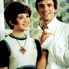 Zwei Menschen lächeln, der Mann trägt ein weißes Hemd, die Frau ein Kleid mit großem Medaillon, sie halten sich an den Händen.