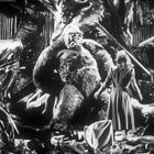 Eine Frau steht vor einem großen Gorilla inmitten von Trümmern, beide blicken sich an.