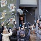 Menschenmenge mit Kameras und Mikrofonen vor einem Mann in einer Tür. Plakate an der Wand: "Grüne Hochzeit", "Treffen im Bureaü".