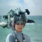 Eine Person mit außergewöhnlicher Frisur aus Materialien vor einem verschwommenen Meereshintergrund.
