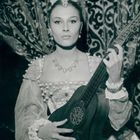 Eine Frau in historischer Kleidung hält eine Laute vor einem gemusterten Vorhang. Sie trägt Schmuck und schaut ernst.