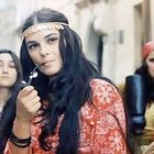 Mehrere Frauen in bunter Kleidung, eine raucht Pfeife, trägt Blumenstirnband, andere schauen ernst, Hintergrund ist unscharf.