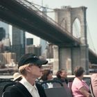 Keine sichtbaren Texte. Menschen auf einer Fähre in der Stadt, im Hintergrund Brooklyn Bridge und Wolkenkratzer.