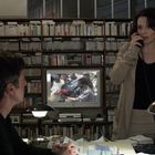 Im Hintergrund läuft ein Fernseher mit unscharfen Nachrichten. Eine Frau telefoniert stehend, ein Mann sitzt und schaut zu. Bücherwand im Hintergrund.