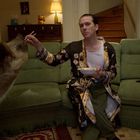 Ein Mann im gemusterten Mantel sitzt auf einem grünen Sofa, hält Essstäbchen und teilt Essen mit einem großen Hund.