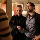 Eine Frau und ein Mann stehen nebeneinander, sie hält einen Teller. Beide schauen ernst, im Hintergrund Vasen und Gemälde.