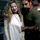 Ein Mann umarmt eine Frau in weißem Kostüm mit aufgemaltem Gesicht. Im Hintergrund ein Kamin mit Deko und Bücherregale.