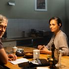 Ein Mann und eine Frau sitzen in einer Küche. Auf dem Tisch liegen Papiere, eine Tasse und eine Schale.