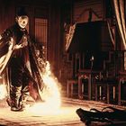 Eine Person in traditioneller Kleidung steht in einem Raum, umgeben von Flammen, umgeben von alten Holzmöbeln.