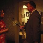 Ich kann die Texte im Bild nicht erkennen. Ein Mann hält eine Frau in einem blumigen Kleid einen kleinen Blumenstrauß hin.