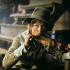 Person mit Hut isst Fleisch in rustikaler Umgebung; Holzstufen im Hintergrund.