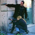 Zahlen "582". Ein Mann steht wachsam, eine Frau kniet mit Gewehr. Beide zielen, versteckt hinter einer Ecke. Spannungsvoll.